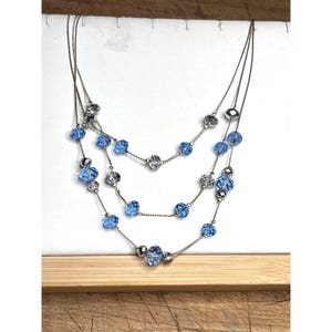May include: A multi-strand necklace with silver-toned chains and blue and clear crystal beads. The necklace has three layers, with the beads arranged in a cascading pattern. The crystals are faceted, catching the light.