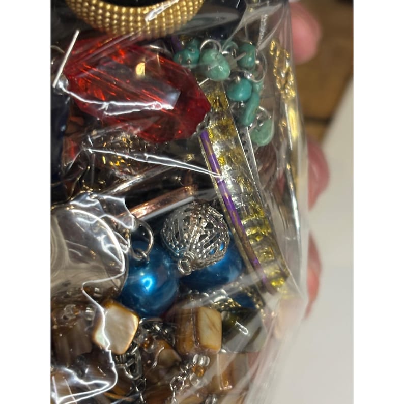 May include: A close-up of a plastic bag filled with a variety of colorful jewelry pieces, including turquoise beads, a red crystal, and a silver charm. The jewelry is arranged in a random pattern and appears to be vintage or antique.