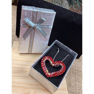 May include: A silver gift box with a light blue ribbon and a heart-shaped necklace with red and clear crystals. The necklace is displayed in an open silver box with a black interior, set on a wooden surface.