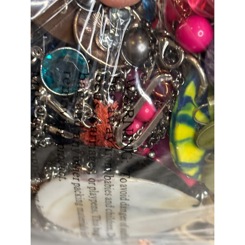 May include: A close-up of a variety of colorful jewelry and trinkets, including beads, charms, and a shell. The jewelry is packaged in a clear plastic bag with a warning label that reads "WARNING: To avoid danger keep away from babies and children. Do not use carmages or playpens. This product is not a toy. Any other packing material should be removed before giving this product to a child."