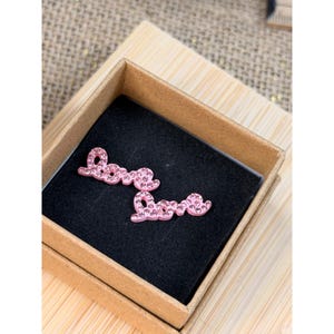 May include: A pair of pink "Love" earrings in a small, open, brown box with a black interior. The earrings are in a cursive font and are encrusted with small, sparkling embellishments.