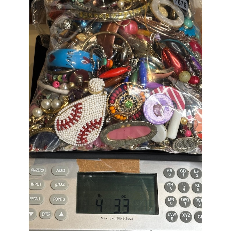 May include: A plastic bag filled with a variety of colorful jewelry and trinkets, including beads, earrings, pendants, and charms. The bag is sitting on a digital scale that reads 4.33 ounces.
