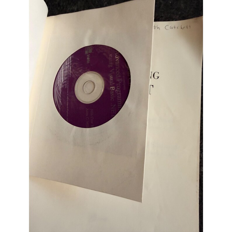 Op de afbeelding: Een paarse cd-rom met de tekst "Advanced Programming using Visual Basic .NET" is zichtbaar in een witte papieren hoes. De hoes zit in een boek met tekst op de rechterpagina.