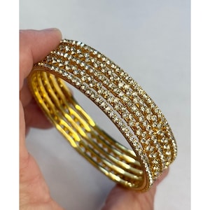 May include: A set of gold bangles, each adorned with multiple rows of sparkling clear crystals. The bangles are held in a hand, showcasing their intricate design and reflective qualities. The gold color provides a warm contrast to the clear crystals.