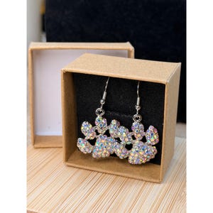 May include: A pair of silver-toned dangle earrings shaped like paw prints, encrusted with iridescent rhinestones. The earrings are displayed inside a small, open, brown cardboard box, set against a wooden surface.