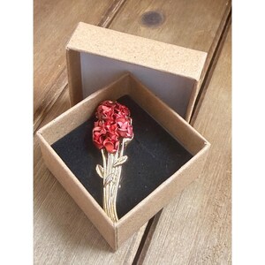 Può includere: Spilla floreale rossa con dettagli color oro, presentata in una piccola scatola quadrata di cartone aperta con interno nero. La spilla presenta un gruppo di fiori rossi e foglie verdi.
