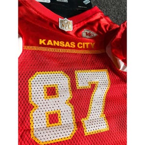 May include: A red mesh jersey with the words "KANSAS CITY" in yellow lettering. The number "87" is prominently displayed in white with yellow trim. An NFL logo is visible.