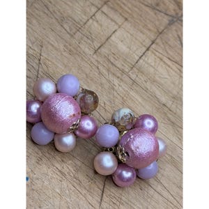 May include: Two vintage clip-on earrings. Each earring features a cluster of various sized beads in shades of pink, lavender, and clear. The earrings have gold-colored metal accents and are set against a wooden surface.