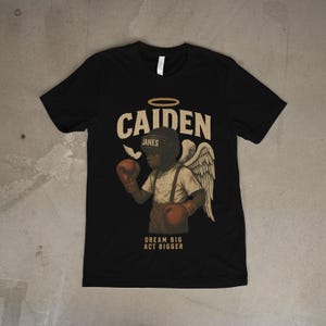 Peut inclure: T-shirt noir avec un graphisme d'un enfant boxeur avec des ailes d'ange, des gants de boxe et un casque. Le texte "CAIDEN JAMES" est au-dessus de l'image, avec "DREAM BIG ACT BIGGER" en dessous.