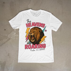 Peut inclure: T-shirt blanc avec un motif de lion rugissant et le texte "The Heavens are Roaring" en rose et noir. Le design comprend des éclairs jaunes et une étoile, avec "Psaume 19:1" en dessous.