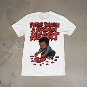 Peut inclure: T-shirt blanc avec un graphisme d'un enfant essayant de reconstituer un cœur brisé. Le texte "Piecing Together A Broken Heart" est imprimé en rouge au-dessus de l'illustration. L'enfant porte un t-shirt turquoise et un pantalon noir.