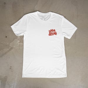 Peut inclure: Un t-shirt blanc à col rond et manches courtes. Le texte rouge "OPEN DOORS" est imprimé sur le côté supérieur gauche de la poitrine. Le t-shirt est fait d'une matière douce et légère, posé à plat sur une surface neutre.