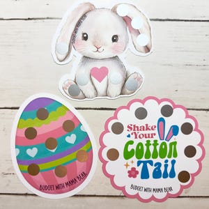 May include: Three Easter-themed stickers on a white wooden surface. The stickers include a white bunny with a pink heart, an Easter egg with colorful stripes and hearts, and a pink scalloped circle with the text "Shake Your Cotton Tail".