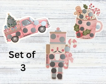 May include: A set of three Christmas-themed scratch-off stickers featuring a pink pickup truck carrying a tree, a mug of hot cocoa with a gingerbread man, and a nutcracker. Each sticker has circular silver scratch-off areas and the text Set of 3.