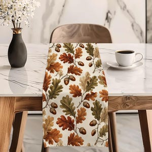 Op de afbeelding: Een rechthoekige tafelloper met een herhalend patroon van bruine eikels en eikenbladeren in tinten groen, oranje en beige op een crèmekleurige achtergrond. De stof toont gedetailleerde botanische illustraties van herfstbladeren en noten.