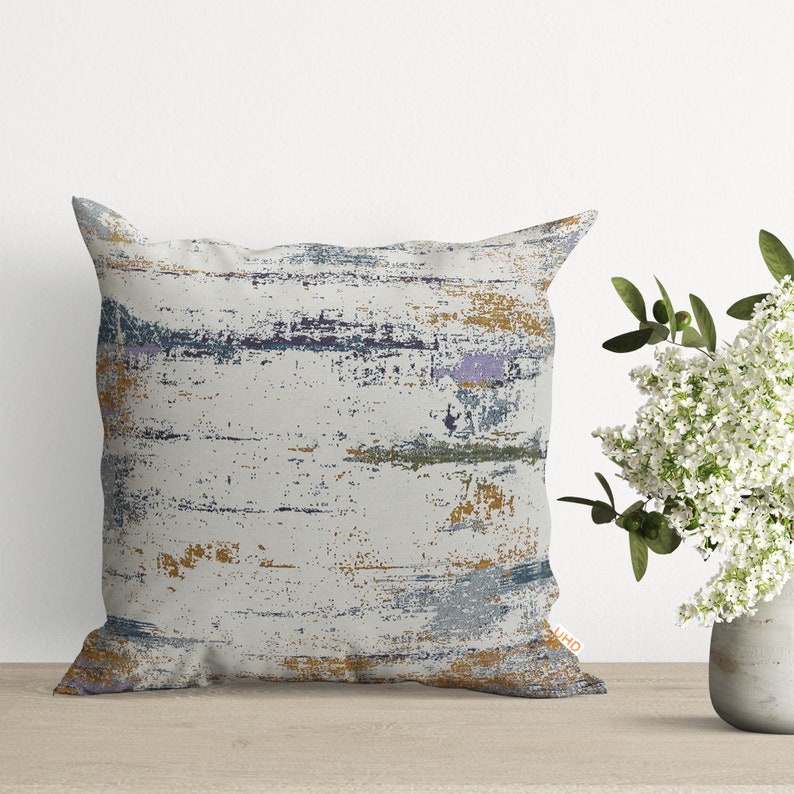 Pu&ograve; includere: Un cuscino decorativo con sfondo bianco e un motivo astratto in tonalit&agrave; di blu, viola e oro. Il motivo ricorda pennellate o legno invecchiato.