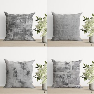 Op de afbeelding: Vier grijze kussens met een verweerd, abstract patroon. De kussens zijn gerangschikt in twee rijen van twee, met een witte bloemenarrangement voor elk kussen.