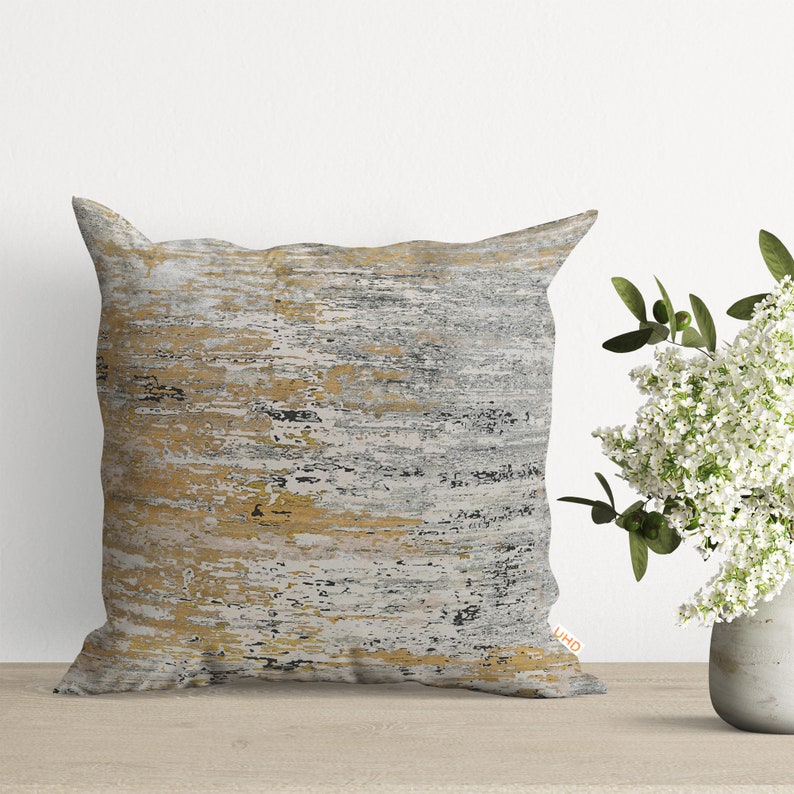 Pu&ograve; includere: Un cuscino decorativo con un motivo astratto grigio, oro e bianco. Il cuscino &egrave; su una superficie di legno.