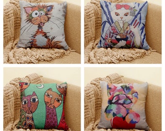 May include: Four decorative throw pillows with different cat designs. The pillows are on a light brown couch with a light brown fuzzy throw blanket. The first pillow features a cartoon cat with big eyes and a surprised expression. The second pillow features a cat wearing a kimono and holding chopsticks. The third pillow features three cats with colorful patterns. The fourth pillow features a cat with a colorful abstract design.