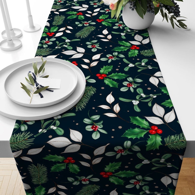 A navy blue table runner with a repeating pattern of green holly leaves, red berries, and white sprigs of mistletoe. The pattern is set against a dark blue background with small white dots.
