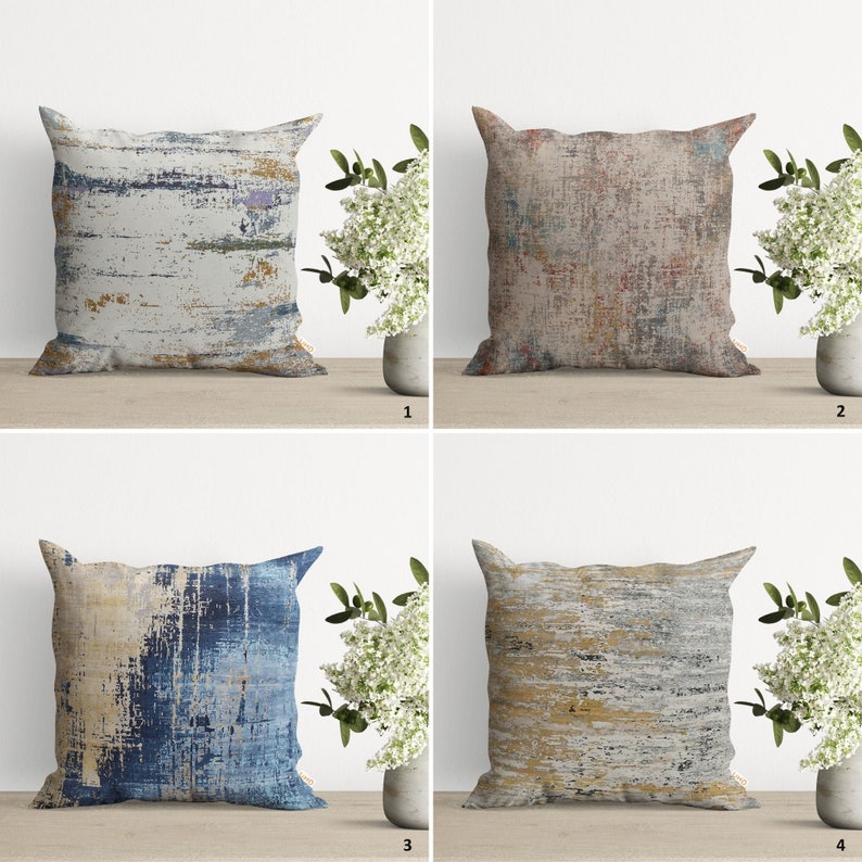Pu&ograve; includere: Quattro cuscini decorativi con motivi astratti in tonalit&agrave; di blu, marrone, giallo e grigio. I cuscini sono disposti in una griglia su una superficie in legno.