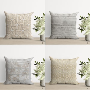 Op de afbeelding: Vier decoratieve sierkussens met verschillende geometrische en textuurontwerpen. De kussens zijn vierkant en hebben patronen in goud, grijs en wit. Ze zijn gerangschikt op een licht oppervlak, vergezeld van een vaas met witte bloemen.