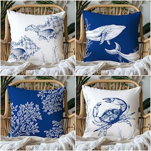 Może przedstawiać: Cztery dekoracyjne poduszki z motywami marynistycznymi. Poduszki przedstawiają białe i granatowe ilustracje muszli, wieloryba, koralowców i kraba. Poduszki są kwadratowe i umieszczone na wiklinowym krześle z białym kocem.
