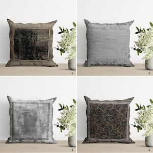 Op de afbeelding: Vier decoratieve kussens met een geometrische rand en verschillende textuur stoffen inzetstukken. De kussens zijn gerangschikt in een raster op een houten oppervlak. De kussens zijn in tinten grijs, zwart en goud.