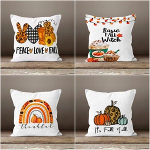 May include: Four white throw pillows with fall-themed designs. The first pillow has a peace sign, a heart, and the words "Peace Love Fall". The second pillow has a coffee cup, a cupcake, and the words "Basic Fall Witch". The third pillow has a rainbow with pumpkins and the word "thankful". The fourth pillow has pumpkins and the words "It's Fall Y'all".