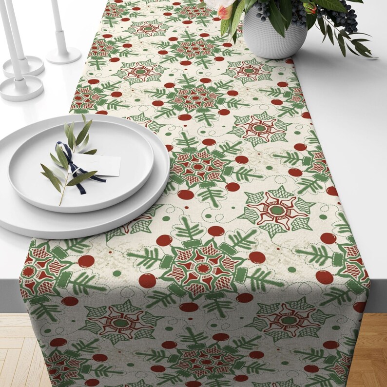 May include: A white table runner with a green and red snowflake pattern. The runner is decorated with small red dots and has a subtle distressed look.