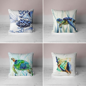 Może przedstawiać: Cztery dekoracyjne poszewki na poduszki w morskim stylu. Poduszki zdobią akwarelowe ilustracje muszli, żółwi morskich i wodorostów. Wszystkie poduszki są białe z niebieskimi i zielonymi akcentami.