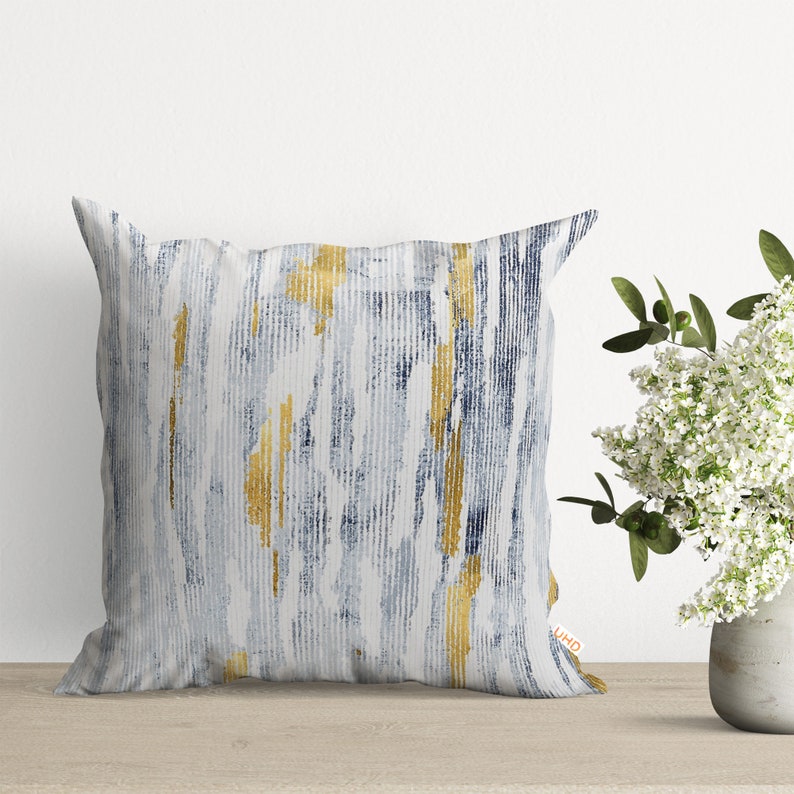 May include: A white throw pillow with a blue and gold abstract pattern. The pattern resembles brushstrokes and stripes.
