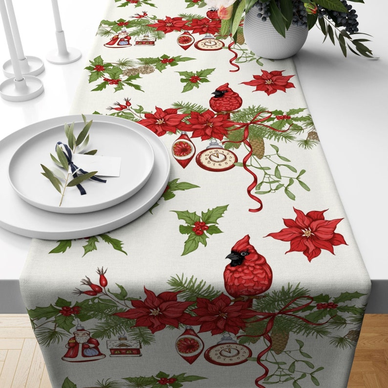 White table runner with a repeating pattern of red poinsettias, cardinal birds, pine branches, holly, and antique clock ornaments.