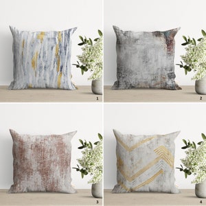 May include: Four decorative throw pillows with different abstract patterns. The pillows are all white with gold, blue, red, and brown accents. The pillows are arranged in a grid pattern on a wooden surface.