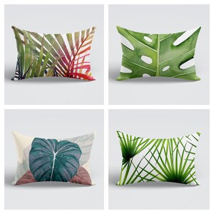 May include: Four different throw pillow designs featuring tropical leaf patterns. The pillows are rectangular and have a white background. The designs include green and red palm leaves, a large green monstera leaf, a large teal leaf, and a cluster of green palm fronds.