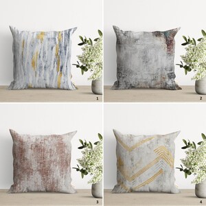 Op de afbeelding: Vier decoratieve kussens met verschillende abstracte patronen. De kussens zijn allemaal vierkant en hebben een witte achtergrond met gouden en grijze accenten. De kussens zijn gerangschikt in een rasterpatroon op een houten oppervlak.