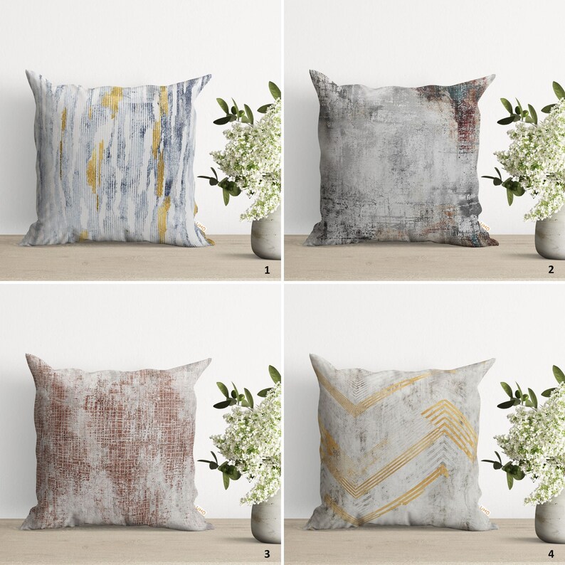 May include: Four decorative throw pillows with different abstract patterns. The pillows are all square and have a white background with gold and gray accents. The pillows are arranged in a grid pattern on a wooden surface.