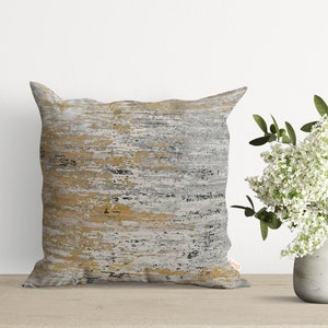 Pu&ograve; includere: Un cuscino decorativo con un motivo astratto grigio, oro e bianco. Il cuscino &egrave; su una superficie di legno.
