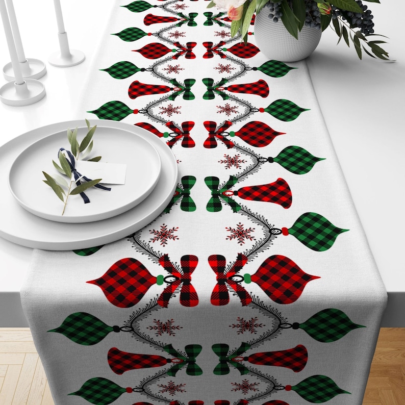 May include: A white table runner with a repeating pattern of red and green plaid ornaments, snowflakes, and bows. The ornaments are hanging from a string of beads.