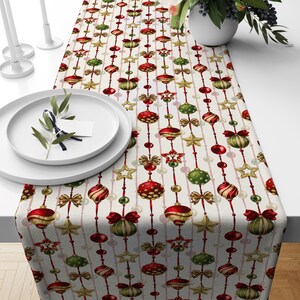May include: A white table runner with a repeating pattern of red, green, and gold Christmas ornaments, stars, and bows. The ornaments are strung together with white beads.