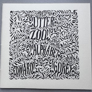 Op de afbeelding: Zwart-wit illustratie van een boekomslag. De titel, "The Gutter Zoo Alphabet by Edward Gorey", is in een decoratief lettertype, omgeven door een bladerenpatroon. Het kunstwerk staat op een getextureerde, gebroken witte achtergrond.