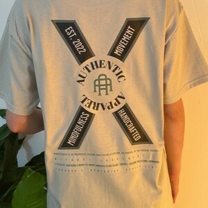 May include: A light gray t-shirt with a black and green logo on the back. The logo says "Authentic Apparel" with a stylized "A" in the center. The logo is surrounded by a circle with the words "Mindfulness" and "Handcrafted" on either side. The words "Est. 2022" and "Movement" are on the top of the logo.
