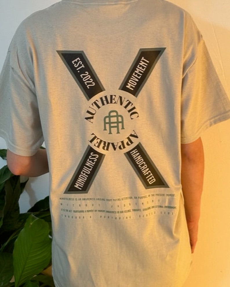 May include: A light gray t-shirt with a black and green logo on the back. The logo says "Authentic Apparel" with a stylized "A" in the center. The logo is surrounded by a circle with the words "Mindfulness" and "Handcrafted" on either side. The words "Est. 2022" and "Movement" are on the top of the logo.