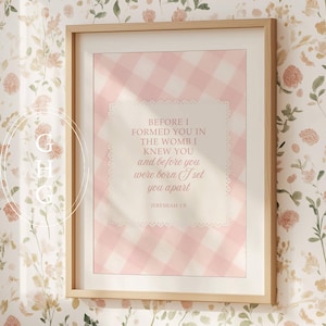 May include: A framed print with a light wood frame. The artwork features a pink gingham pattern background with a white, scalloped-edged overlay containing text from Jeremiah 1:5. The print is displayed against a floral wallpaper.