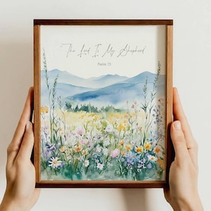 May include: A framed watercolor print featuring a serene landscape with rolling blue mountains and a vibrant meadow of wildflowers. The text "The Lord Is My Shepherd, Psalm 23" is displayed in elegant script. The frame is made of dark wood.