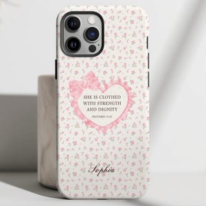 May include: White floral iPhone case with a pink heart-shaped design. The heart contains the text "SHE IS CLOTHED WITH STRENGTH AND DIGNITY" and "PROVERBS 31:25". The name "Sophia" is written at the bottom. The case has a camera cutout.