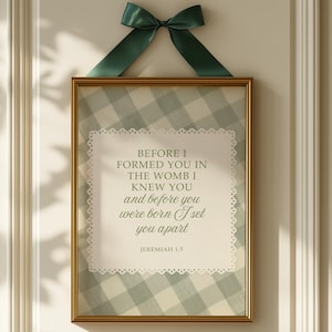 May include: A framed print with a gold-coloured frame, displaying a quote from Jeremiah 1:5. The background has a light blue and white gingham pattern. A dark green ribbon bow is at the top.