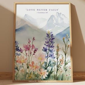 May include: A framed watercolor print featuring a mountain landscape and wildflowers in shades of pink, purple, and orange. The text "LOVE NEVER FAILS" is at the top. The print is in a light-colored wooden frame.