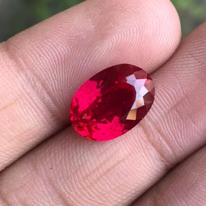 Peut inclure: Une pierre précieuse rouge rubis, de forme ovale et taillée à facettes. La gemme est tenue dans une main, mettant en valeur sa couleur et sa taille. Les facettes de la pierre reflètent la lumière. L'arrière-plan est flou.