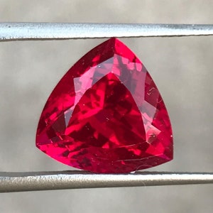 May include: A faceted, triangular-shaped, red gemstone is held by a pair of silver tweezers. The gem has a brilliant cut, reflecting light and showcasing its vibrant color. The background is a blurred, neutral tone.
